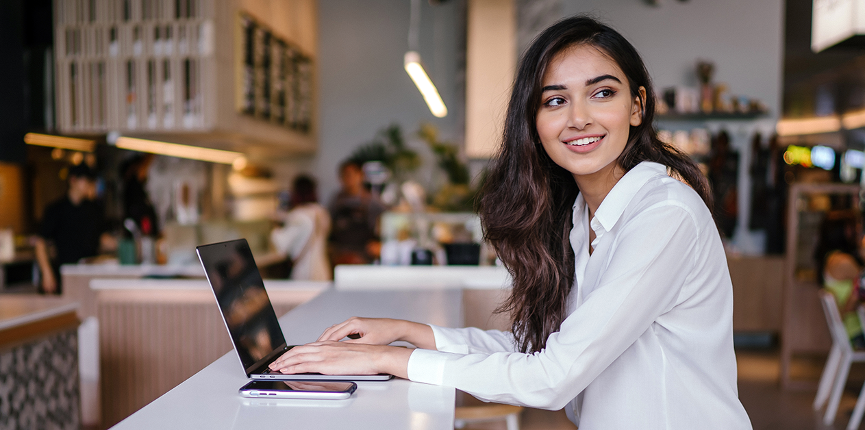 Kvinde arbejder på en bærbar computer på en café og smiler.