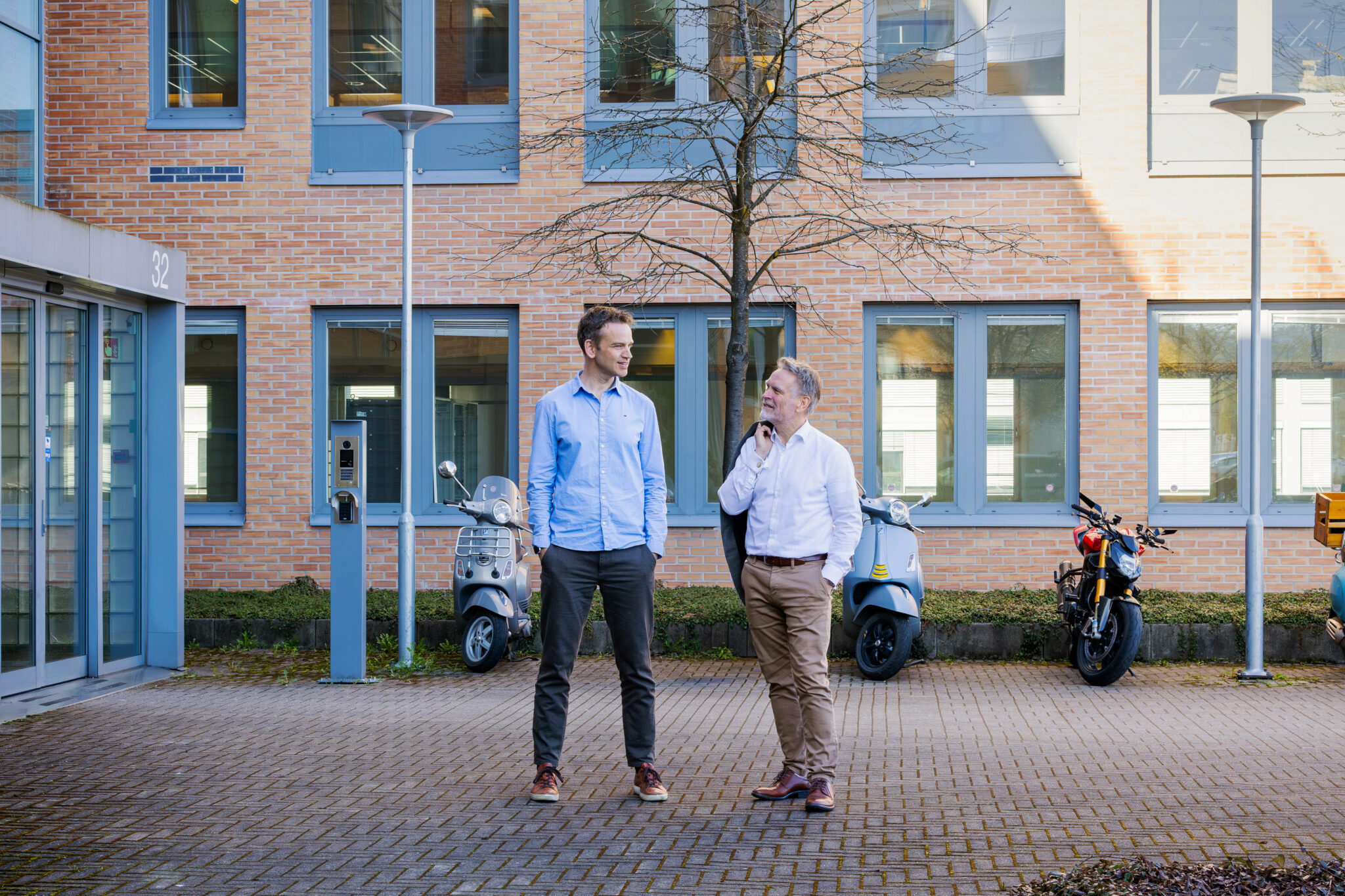 To kolleger st&aring;r utenfor et kontorbygg og snakker sammen, med scootere parkert i bakgrunnen