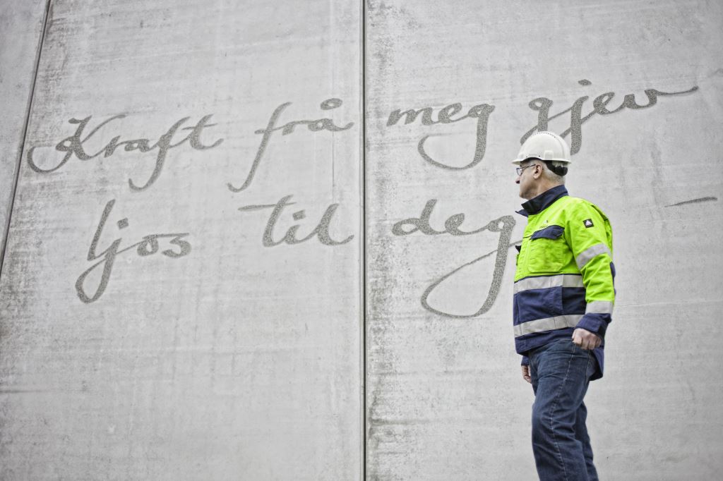 Bilde av en ansatt i Tussa som står i arbeidsklær og ser opp på en grå beting vegg som har teksten "Kraft frå meg gjer ljos til deg"