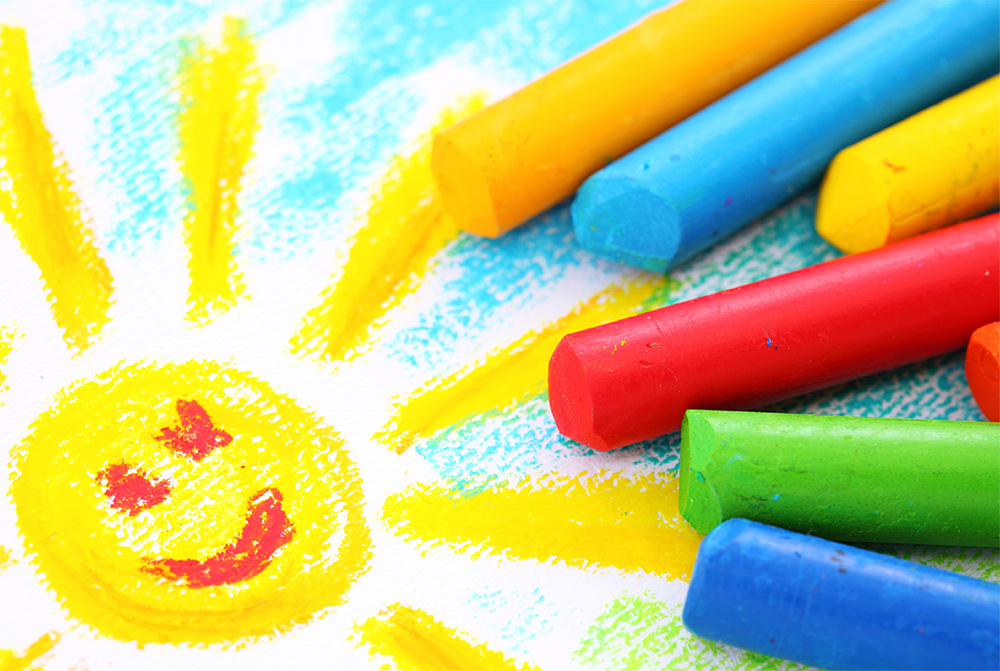 Fargestifter i gul. blå, rød og grønn ligger oppå en barnetegning av en smilende sol med blå himmel