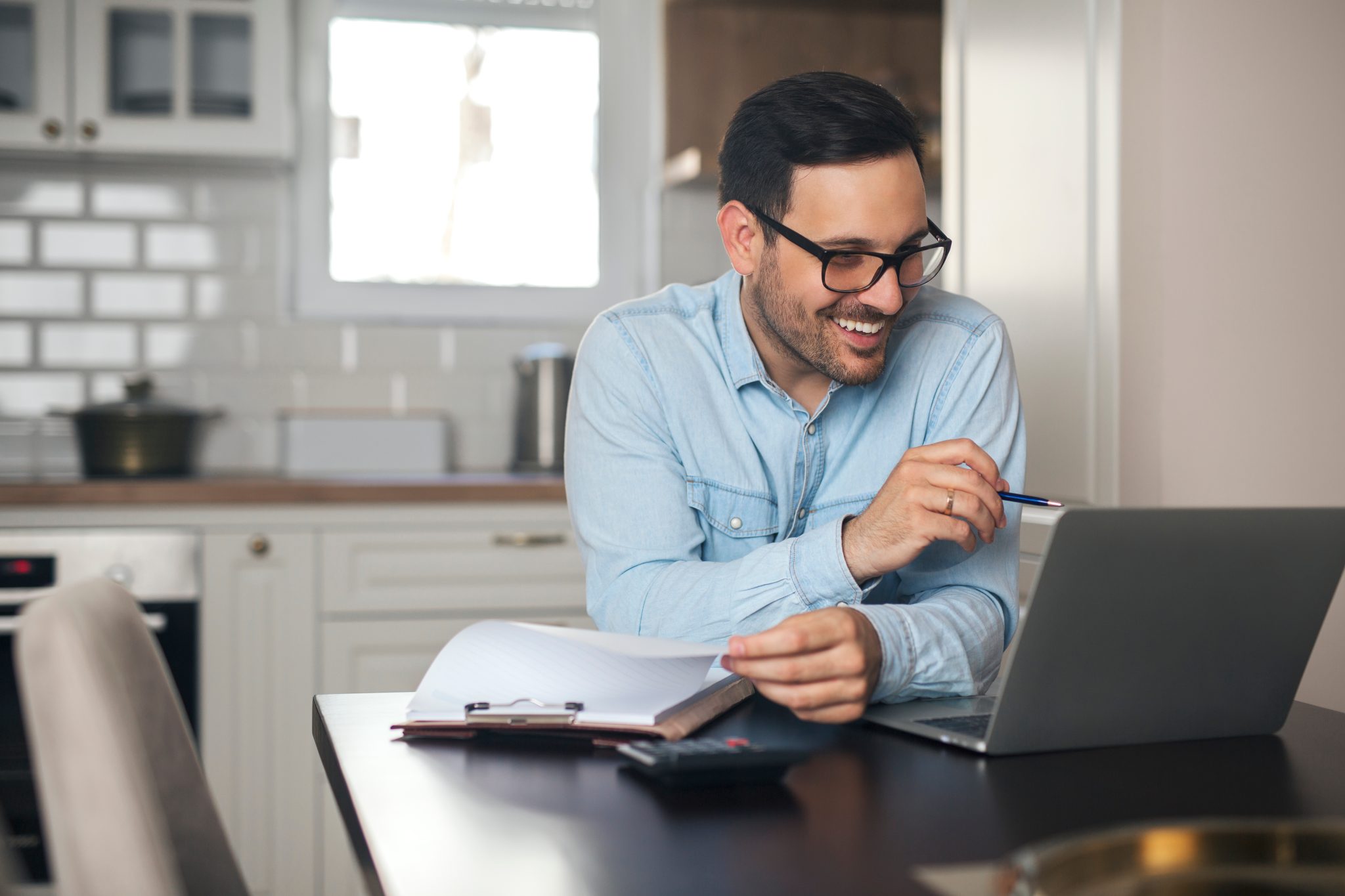 Man working on a laptop holding some paperwork and a pen