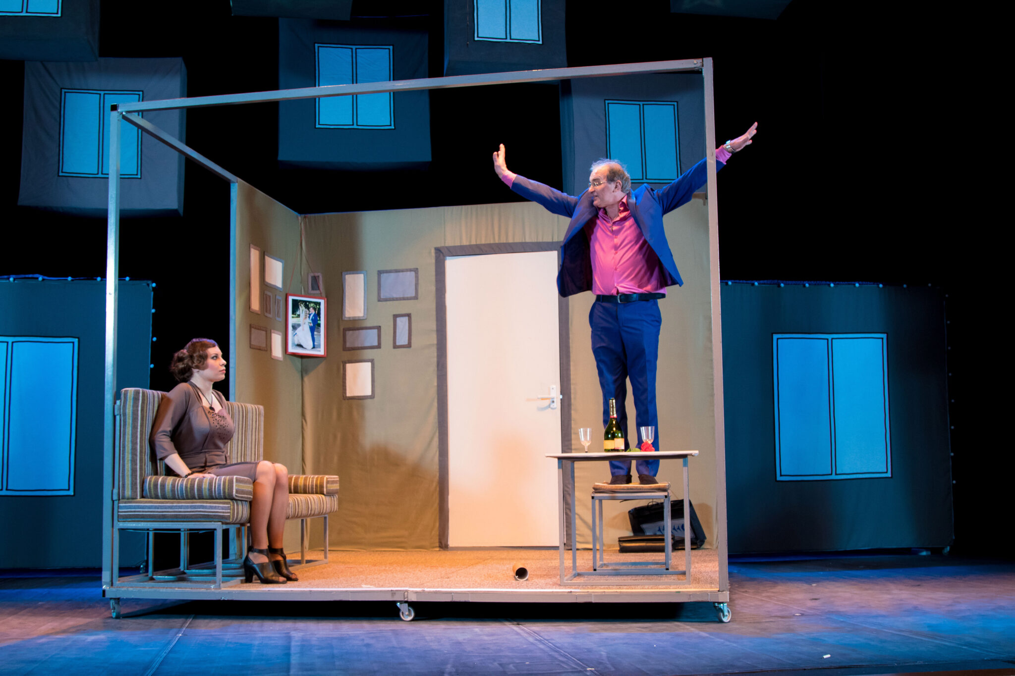 Two actors, brunette woman in a brown dress and a man in a pink shirt and blue suit, playing the role on stage