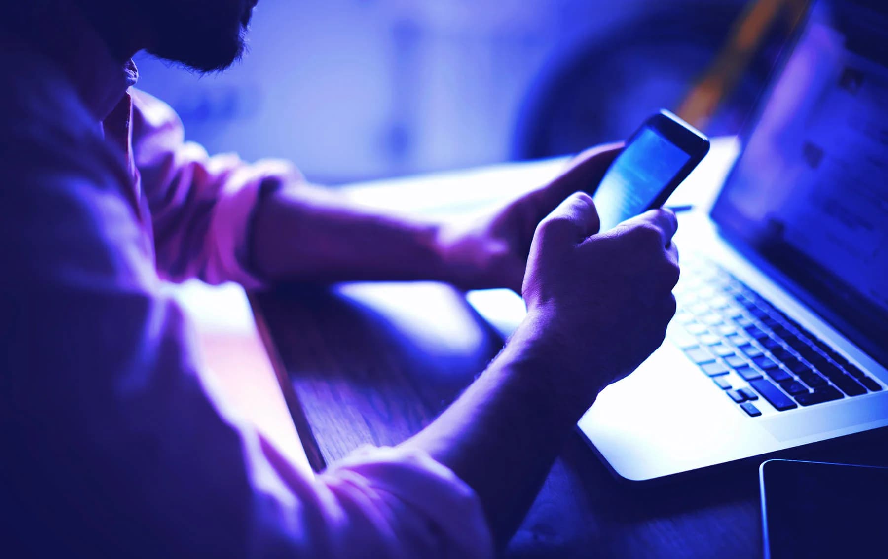 Close-up of a person using a smartphone while sitting at a desk with an open laptop, illuminated by blue and purple lighting.
