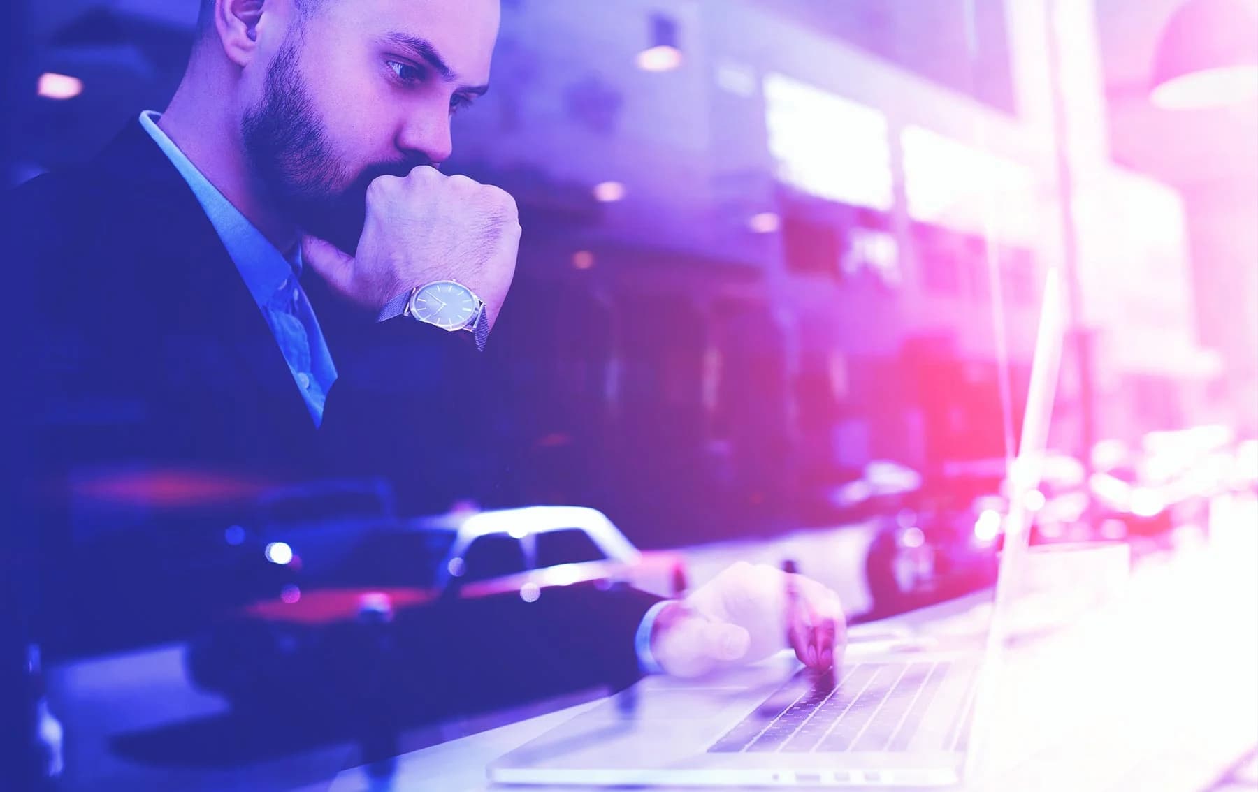 Focused businessman in a suit working on a laptop, resting his chin on his hand, with a city street and cars blurred in the background and purple-blue lighting overlay.