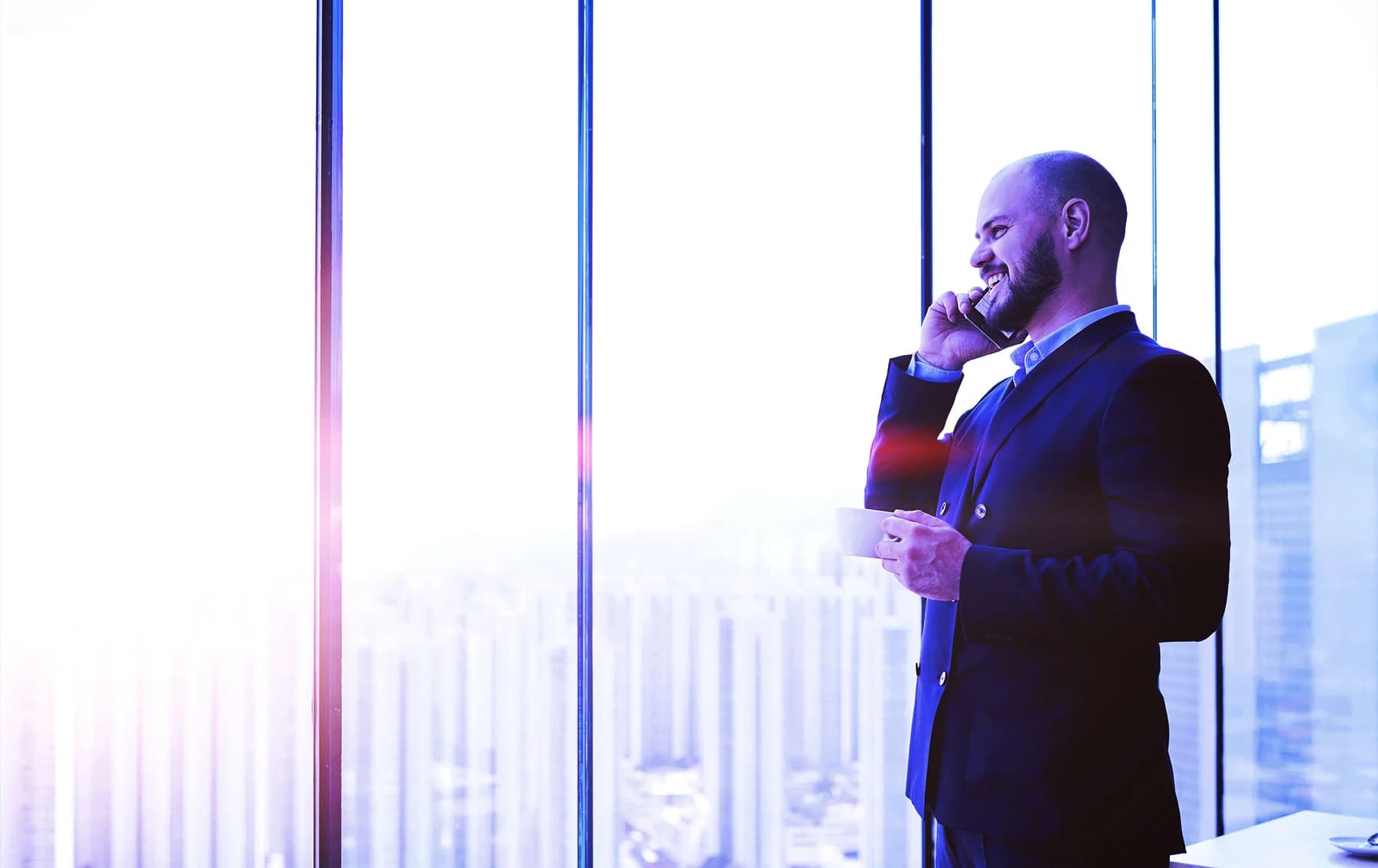 A professionally dressed man in a suit stands by large floor-to-ceiling windows overlooking a city skyline, smiling while talking on his phone and holding a cup, with soft purple and blue lighting effects.
