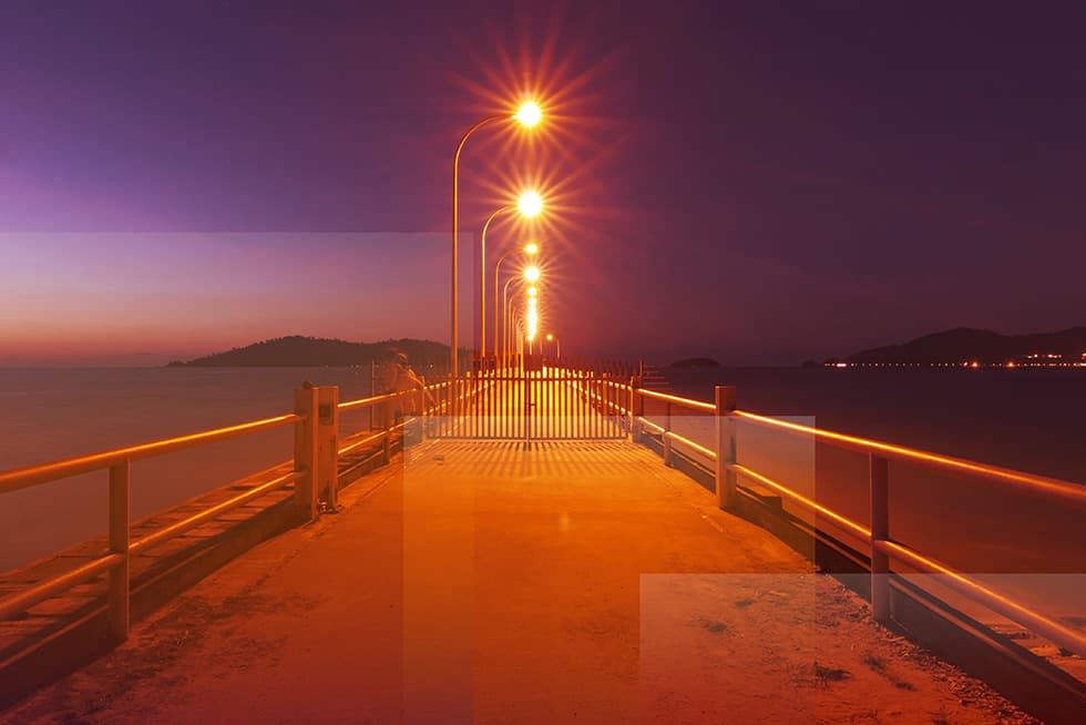 A long pier stretches out over calm water at dusk, illuminated by a row of glowing streetlights that lead toward the horizon.