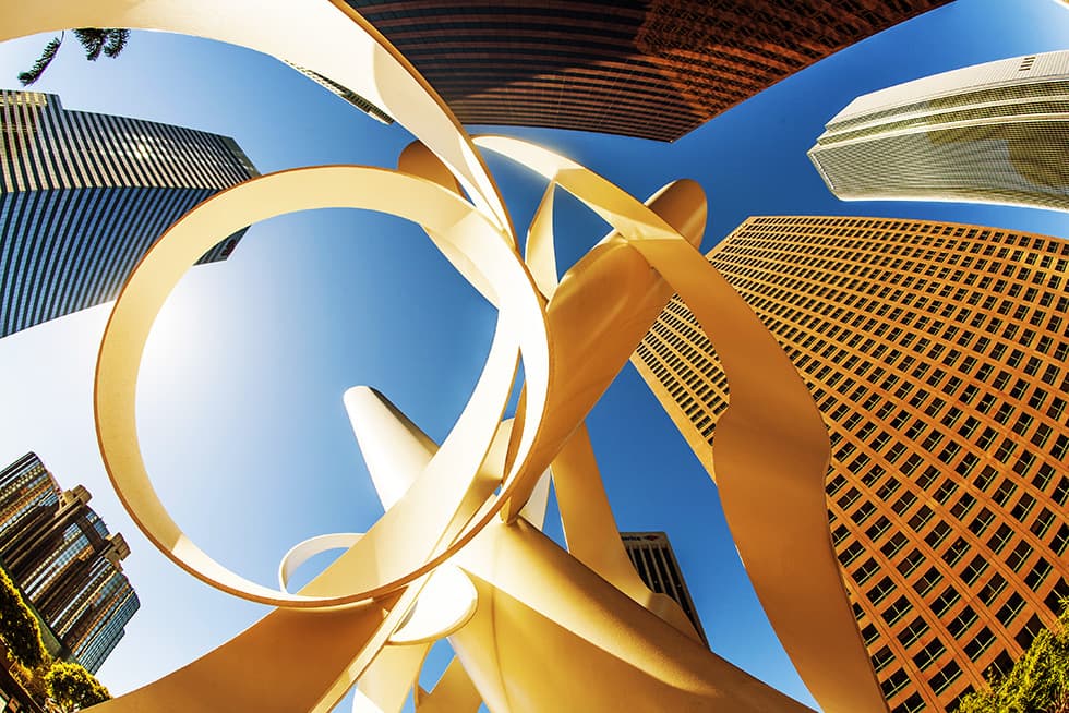 A dramatic upward view of modern skyscrapers surrounding a large abstract metal sculpture, set against a clear blue sky.