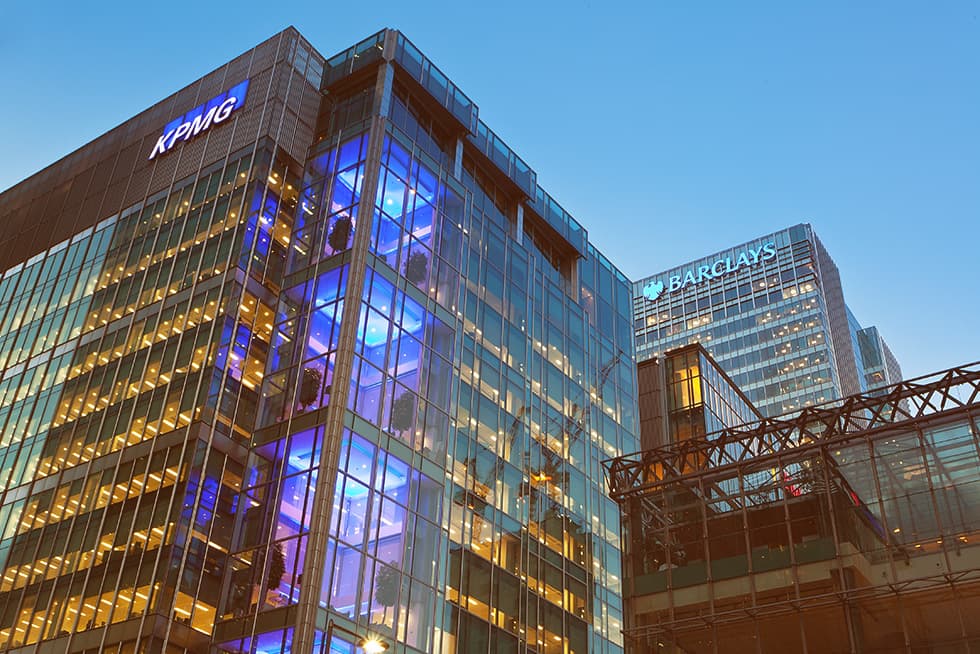 Modern glass office buildings at dusk with illuminated interiors, featuring visible KPMG and Barclays logos on the façades.