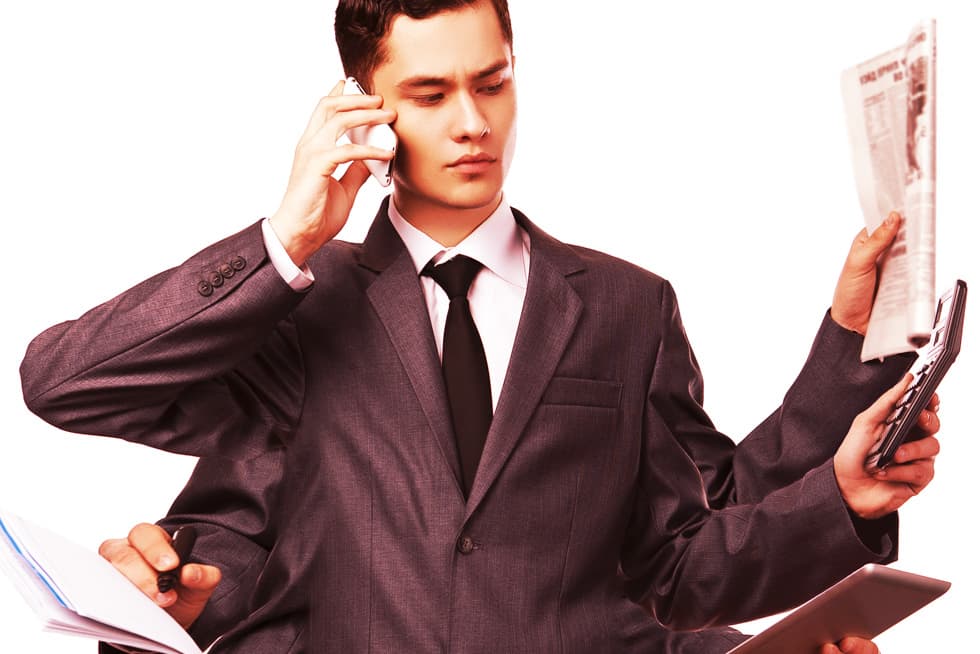 A businessman in a suit appears overwhelmed, holding a phone to his ear while simultaneously managing a newspaper, calculator, documents, and a tablet, suggesting multitasking and heavy workload.