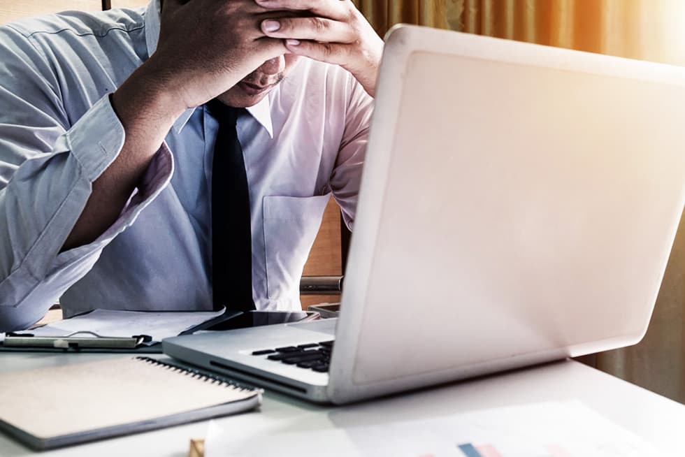 A man in a dress shirt and tie sits at a desk with a laptop, holding his head in his hands, appearing stressed or overwhelmed, with notebooks and papers nearby.