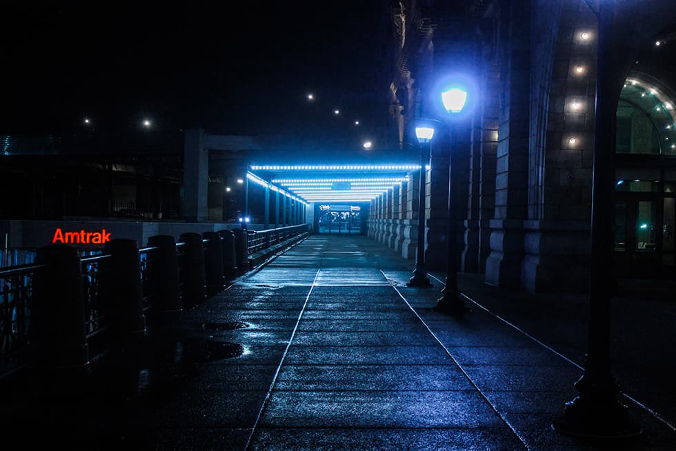 A dimly lit walkway at night with wet pavement reflecting blue overhead lights. Street lamps line the right side beside a large stone building, and a covered, illuminated pathway leads toward glass doors in the distance. A red &ldquo;Amtrak&rdquo; sign glows on the left side near the railing.