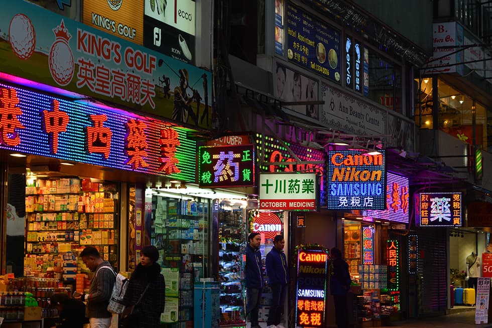 A busy city street at night filled with brightly lit storefronts and colorful neon signs. Many of the signs feature Chinese characters along with brand names like Canon, Nikon, and Samsung. Small shops display goods at their entrances, and several pedestrians walk along the sidewalk under the glow of the illuminated signage.