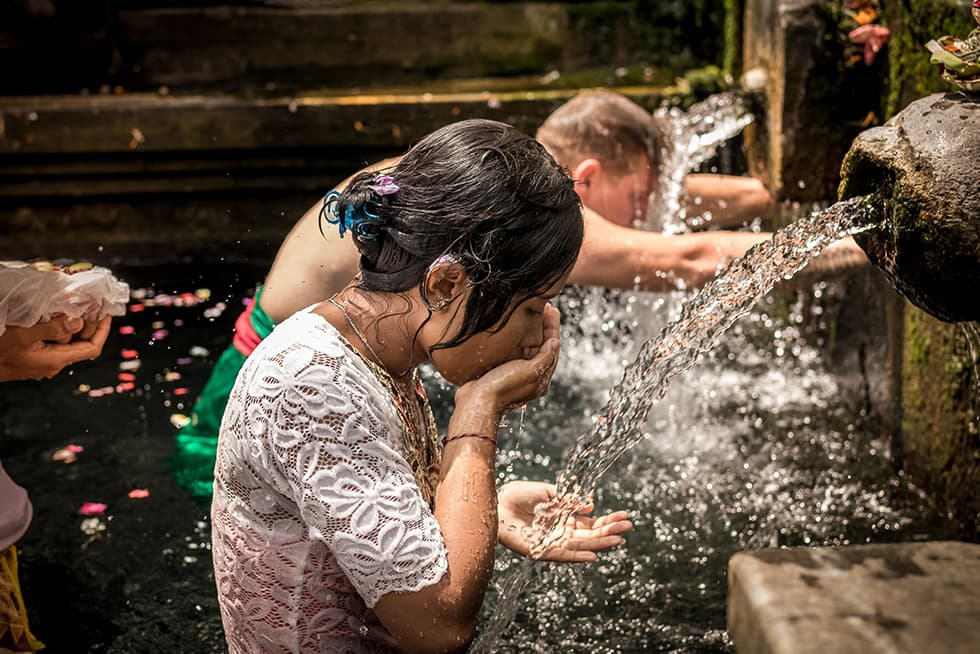 Two children are standing in a shallow stone water feature outdoors. In the foreground, a girl wearing a white lace top cups her hands to drink from a stream of water flowing from a stone spout. Behind her, another child leans forward under a second stream of water. The setting appears natural and serene, with moss-covered stone and flower petals floating on the water&rsquo;s surface.