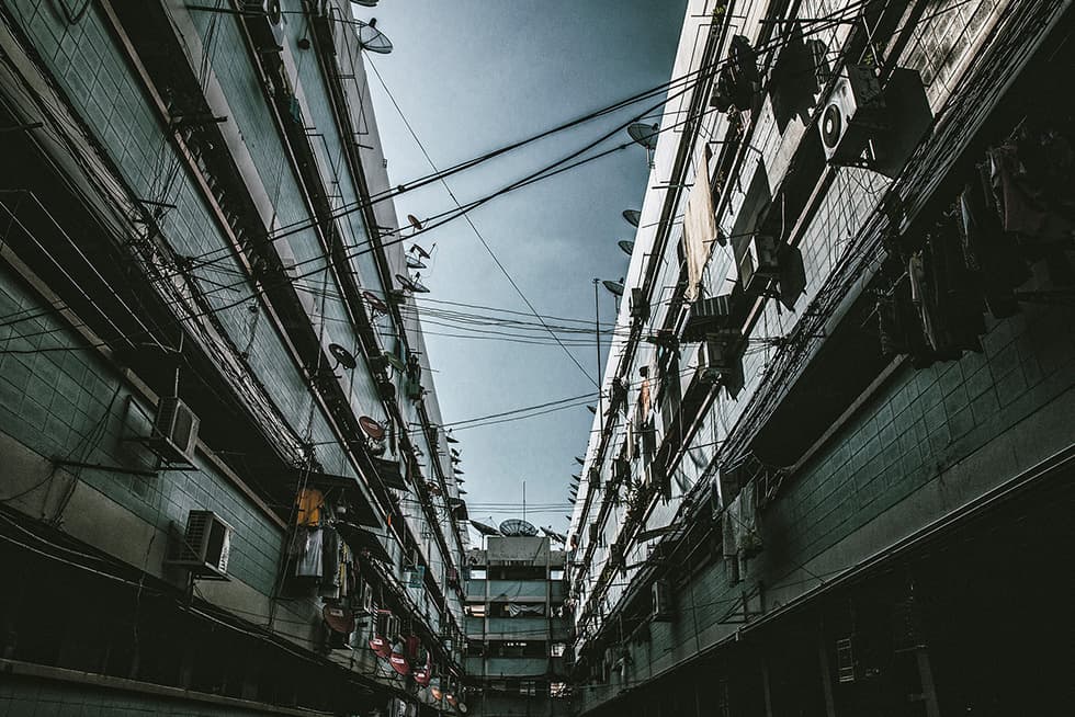 A narrow urban alley framed by two multi-story residential buildings on either side. The buildings have tiled exteriors and are lined with numerous air-conditioning units, satellite dishes, balconies, and hanging laundry. Overhead, a dense web of electrical and communication wires stretches across the gap between the buildings. The sky above is overcast, adding a moody atmosphere to the scene.