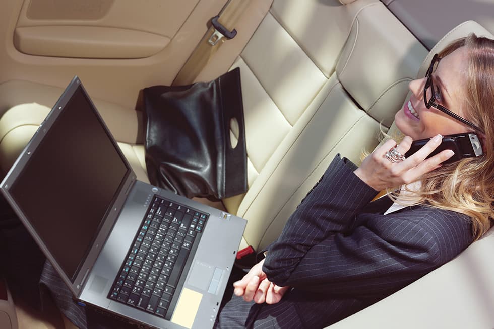 Businesswoman talking on a phone with a laptop while sitting in the back seat of a car.