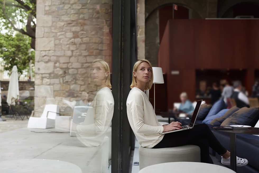 Woman sitting by a window in a caf&eacute; using a laptop, with her reflection visible in the glass.