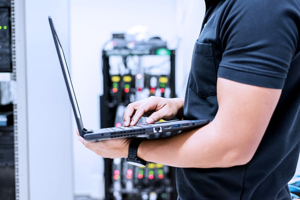 Person using a laptop in a server room with network equipment in the background