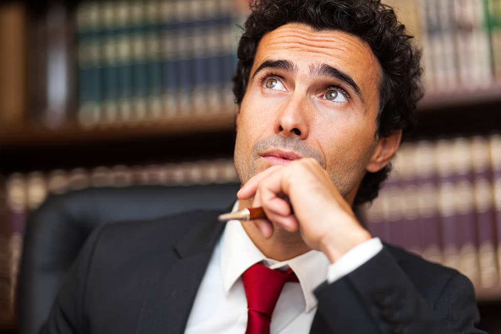 A man in a suit with a red tie sits in front of bookshelves, holding a pen to his chin and looking upward thoughtfully.