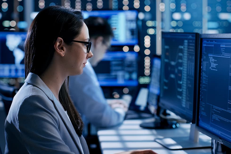 A woman wearing glasses works at a computer in a modern office environment, focusing on code and data displayed on multiple monitors, with another colleague working in the background.