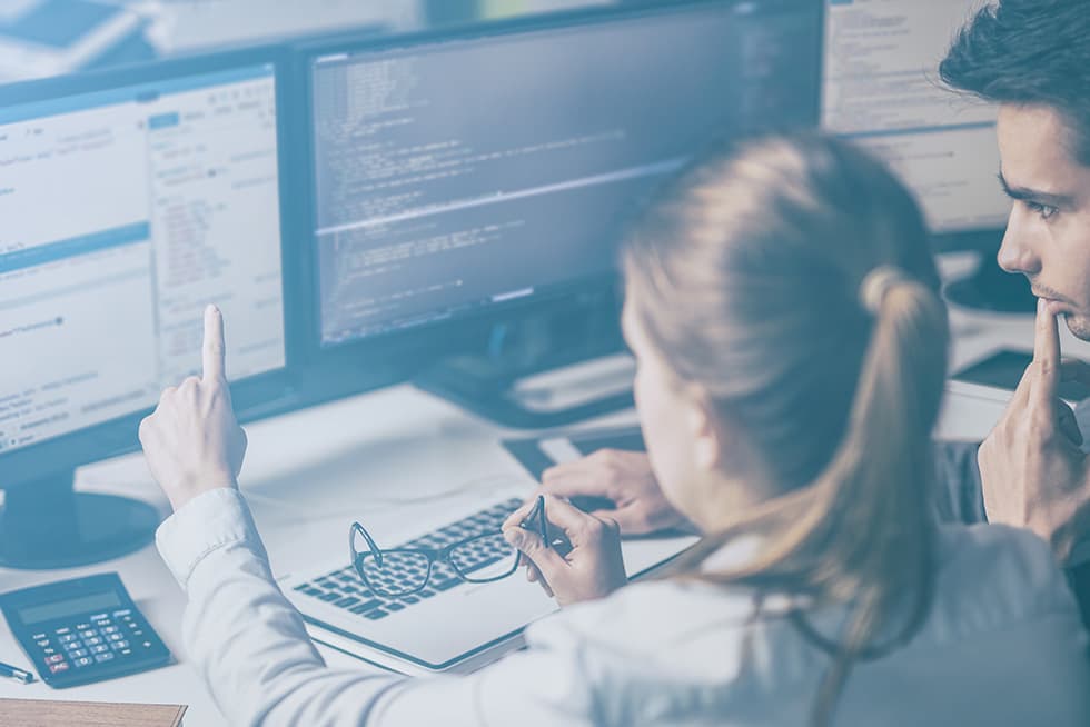 Two colleagues collaborate at a workstation with multiple monitors displaying code, as one points to the screen while discussing the content with the other.