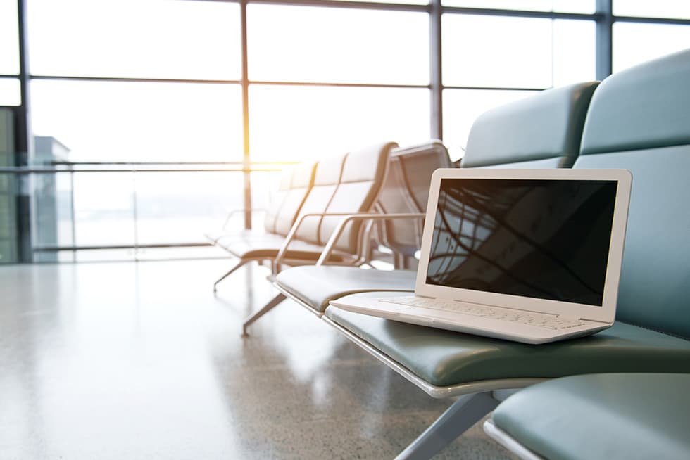 A laptop sits on a row of empty seats in a bright airport terminal waiting area, with large windows and sunlight in the background.
