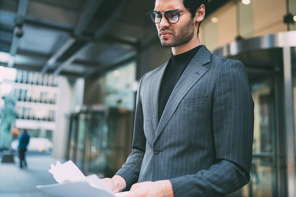 A man in a pinstripe suit and glasses stands outside a modern office building, looking down at documents in his hands with a concerned expression.