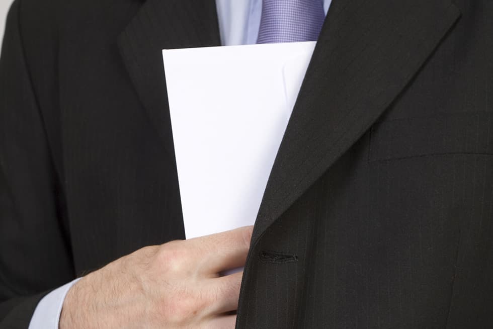 A close-up of a man in a dark business suit holding a white document or folder against his chest.