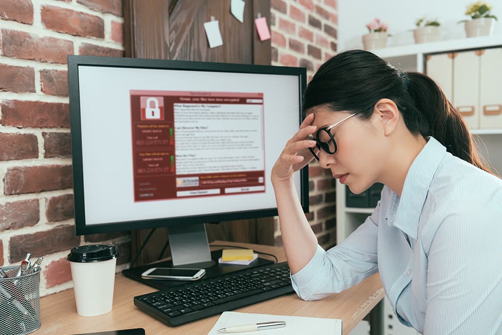 A woman wearing glasses sits at a desk looking stressed, holding her forehead while a computer monitor displays a ransomware warning message with a padlock icon.