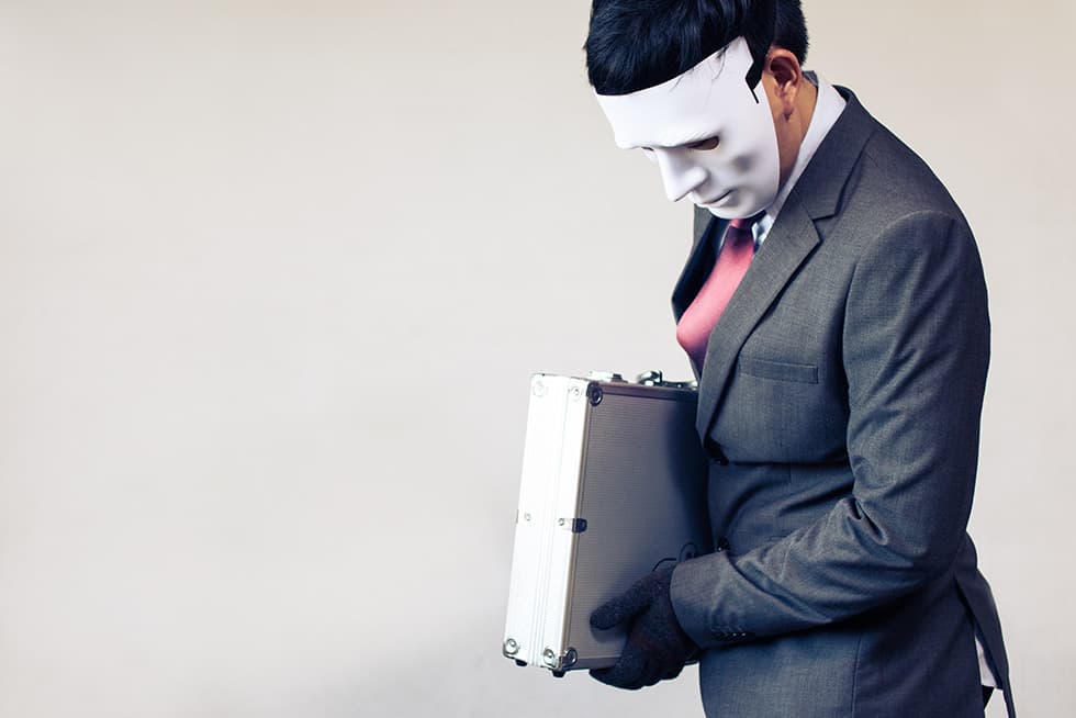 A man in a business suit wearing a white mask and black gloves holds a metal briefcase, looking downward against a plain background.