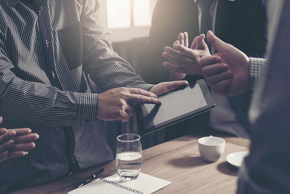 Several business professionals gather around a table, with one person pointing at a tablet screen while others gesture and discuss, and a glass of water and notepad on the table.