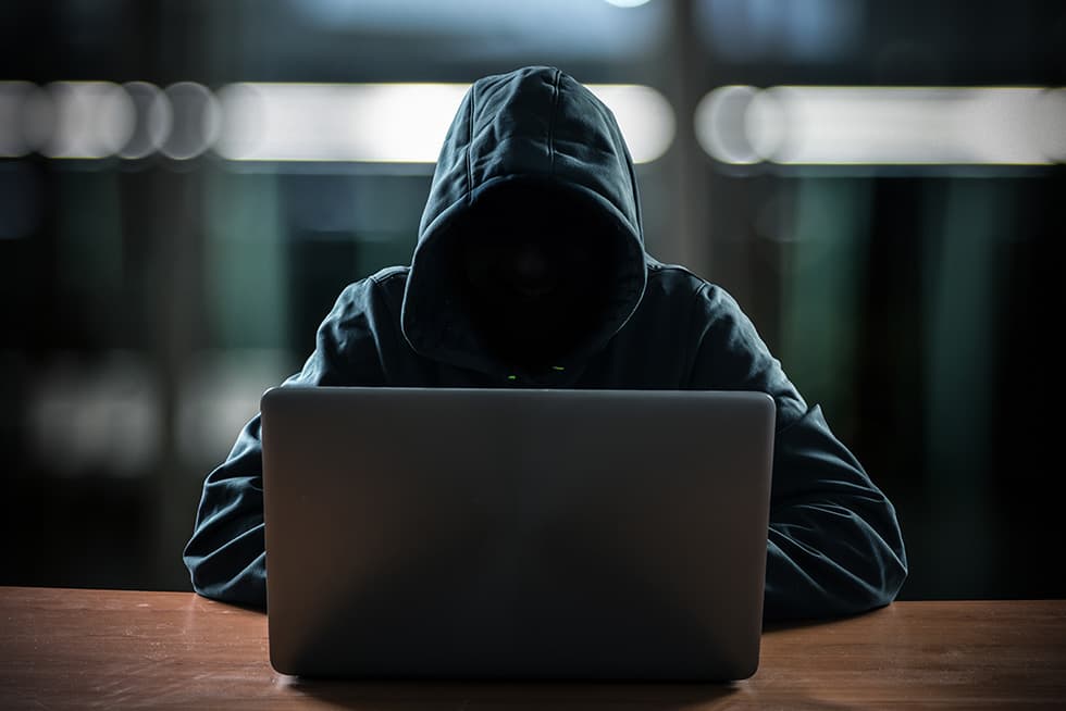 A hooded person sits at a table using a laptop in a dark setting, with their face mostly hidden in shadow.