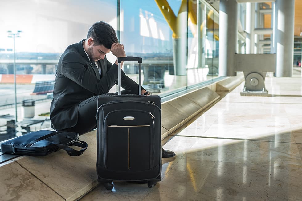 A businessman in a suit sits at an airport terminal beside a rolling suitcase, resting his head on his hand and looking down, appearing stressed or tired.