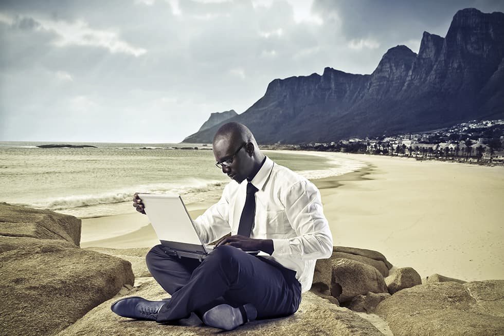 A man in business attire sits on large rocks by the beach, working on a laptop with ocean waves and mountains in the background.