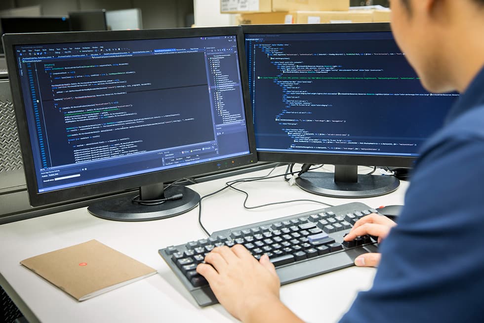 A person types on a keyboard while working at a desk with two monitors displaying lines of computer code.