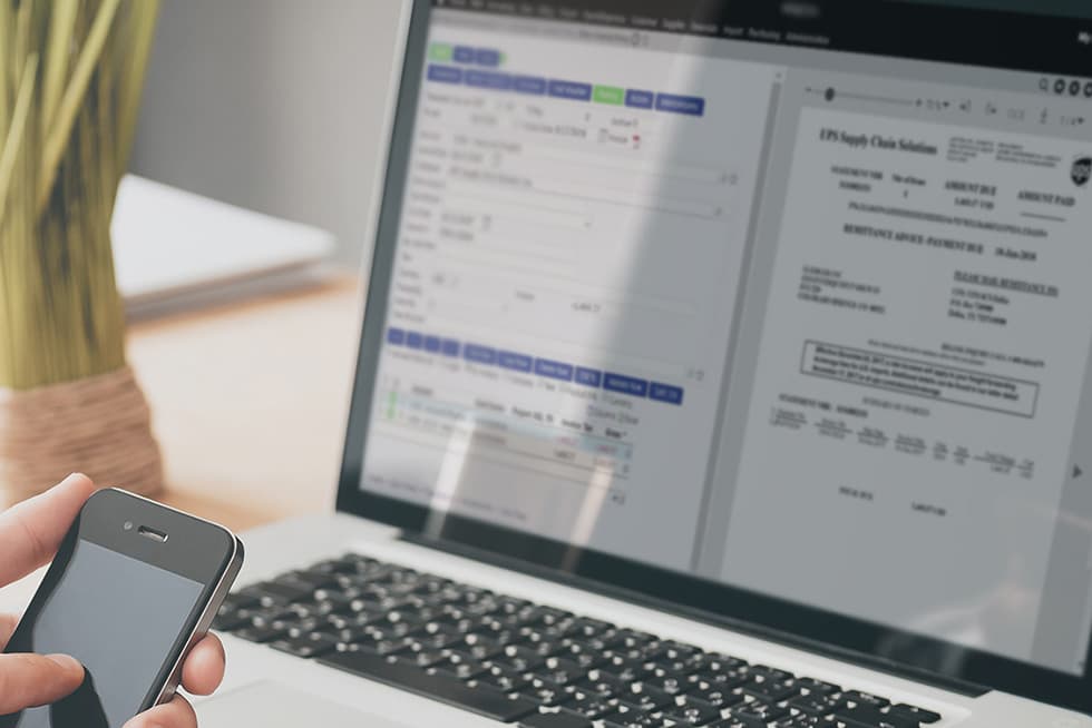 A person holds a smartphone while working on a laptop displaying an invoice or financial document alongside accounting software on the screen.