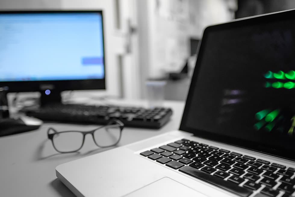 A close-up of a laptop on a desk with a blurred desktop monitor and keyboard in the background, and a pair of eyeglasses resting nearby.