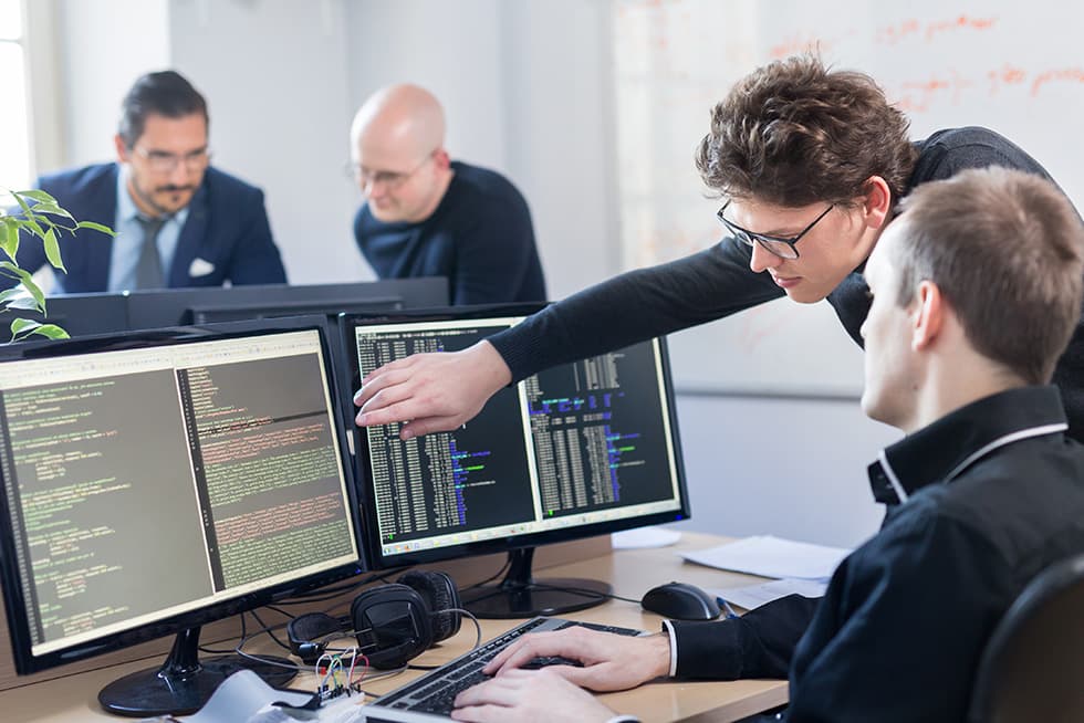 A group of four men working in an office environment, with two seated at dual monitors displaying code while another leans in and points at the screen, and two colleagues discussing in the background.