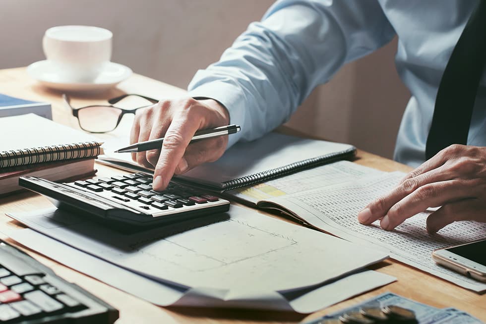 A person in a dress shirt uses a calculator while reviewing financial documents and notes on a desk, with a pen, notebook, and cup of coffee nearby.