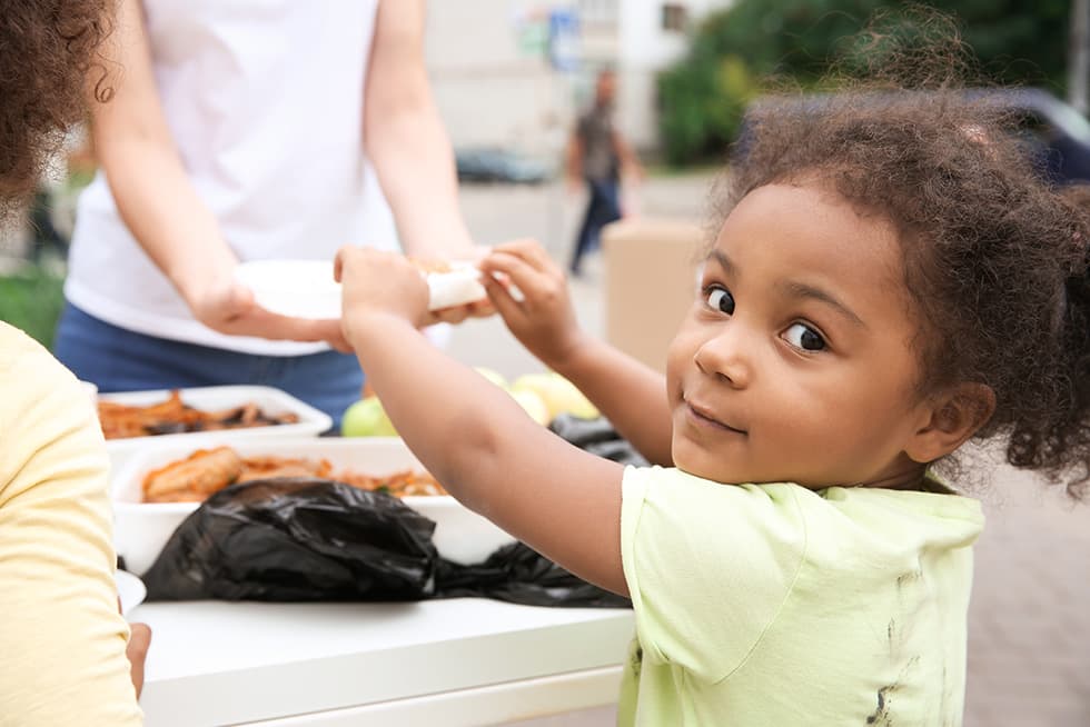 A young child outdoors receives food from an adult across a table with trays of cooked dishes, looking back toward the camera.