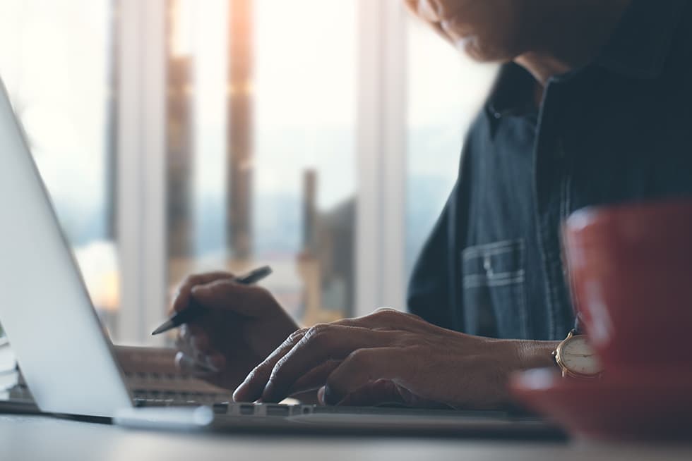 A person works on a laptop near a window, holding a pen while typing, with a cup of coffee in the foreground and warm sunlight in the background.