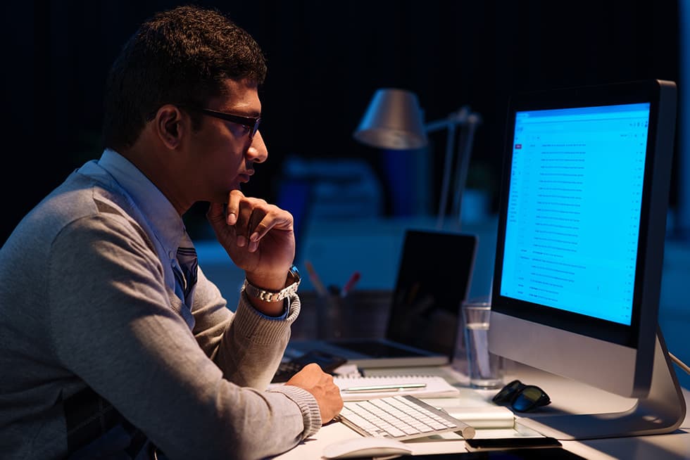 A man wearing glasses sits at a desk at night, looking thoughtfully at a large computer monitor displaying a list of emails, with a keyboard, mouse, and glass of water nearby.
