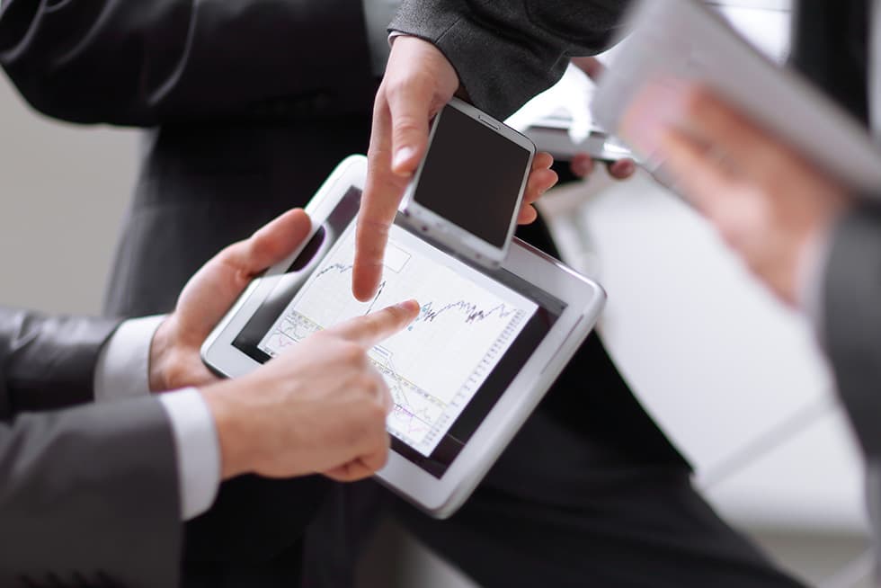 Two business professionals reviewing financial charts on a tablet, with one pointing at the screen while holding a smartphone and documents nearby.