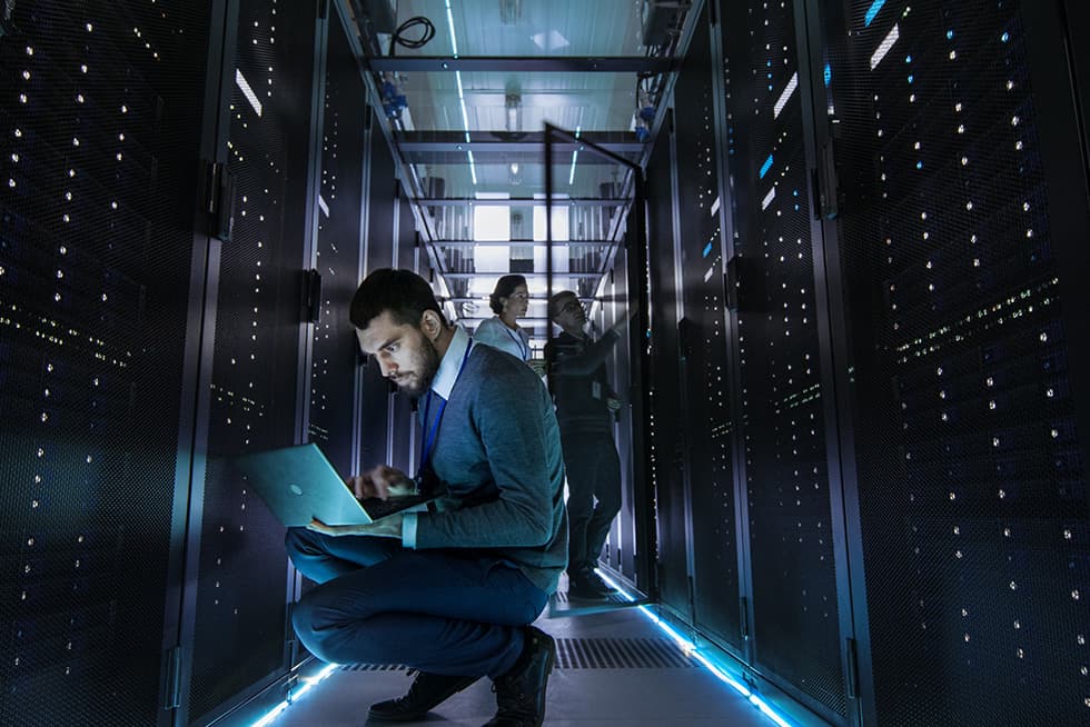 A technician kneels in a data center aisle working on a laptop, surrounded by server racks with illuminated indicators, while two colleagues check equipment in the background.