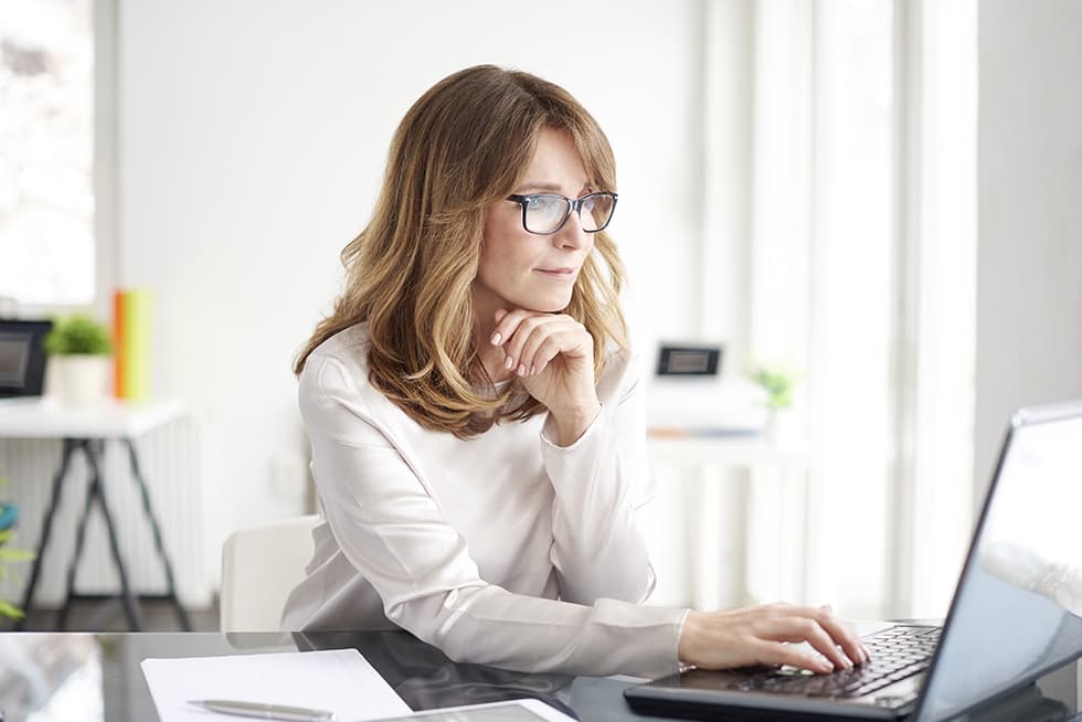 A woman wearing glasses sits at a desk in a bright office, thoughtfully resting her chin on her hand while working on a laptop.
