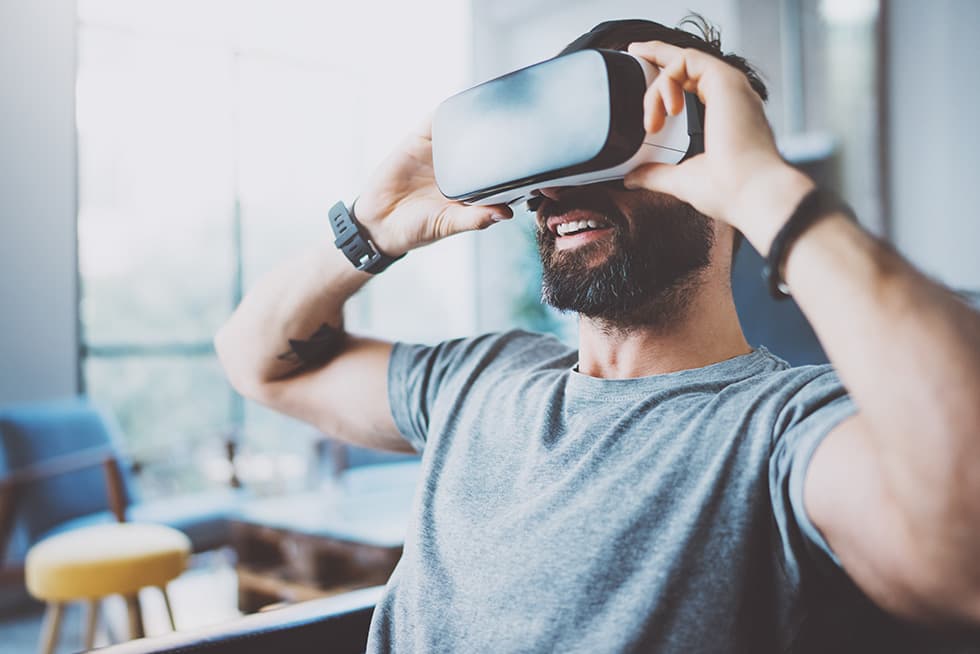A man wearing a virtual reality headset smiles while adjusting it with both hands, sitting indoors in a modern living space.