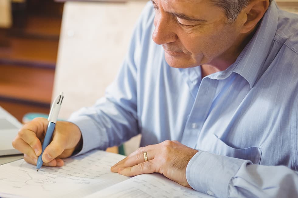 An older man in a light blue shirt writes on a document with a pen, reviewing paperwork at a desk.