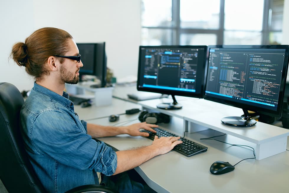 A man with his hair tied back sits at a desk typing on a keyboard while working with two monitors displaying lines of computer code in an office environment.