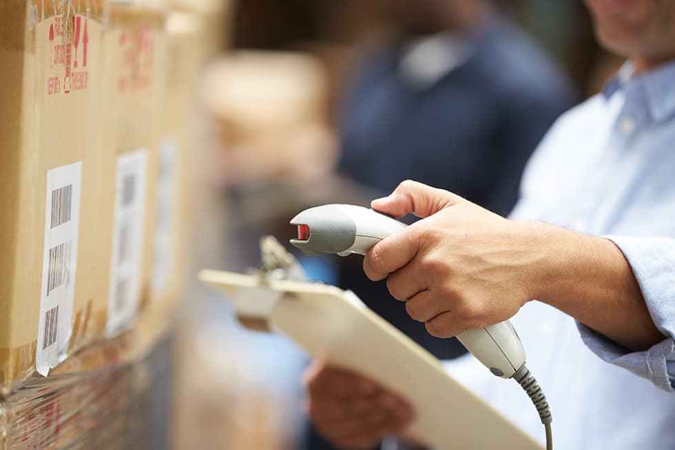 A person uses a handheld barcode scanner to scan labels on stacked cardboard boxes while holding a clipboard.