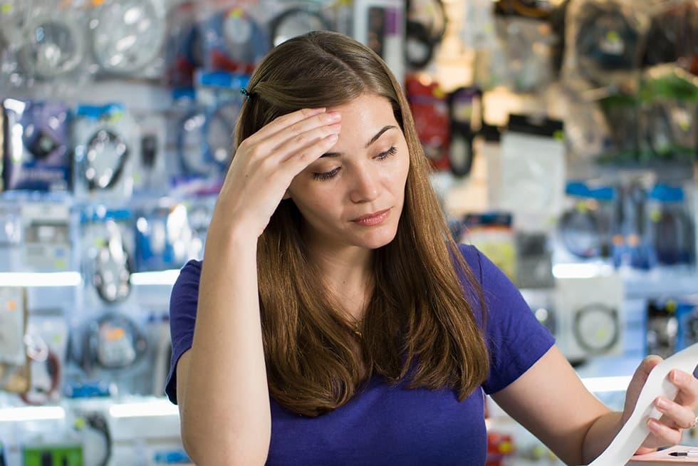 A woman in a store stands in front of shelves filled with packaged electronics accessories, holding a receipt and touching her forehead with a concerned expression.