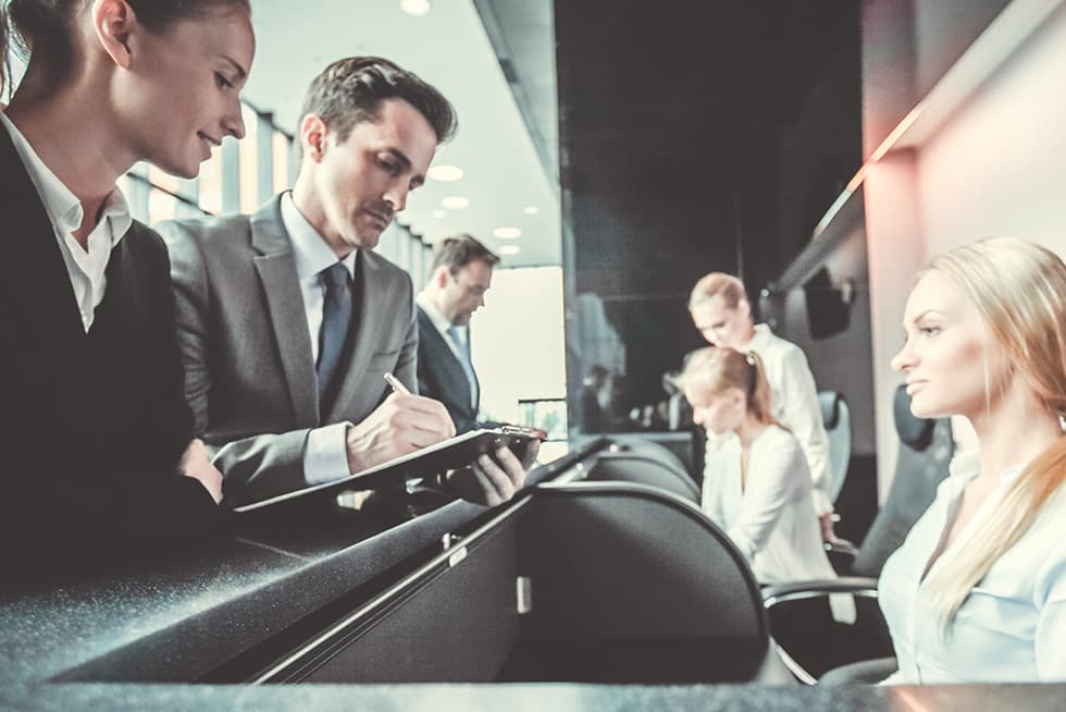 Two professionally dressed employees stand at a reception desk, with one writing on a clipboard while speaking to a seated woman, as other staff members work in the background.